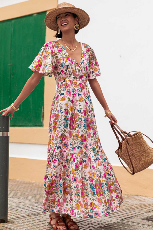 Woman in a floral dress and straw hat holding a bag, standing against a white wall.
